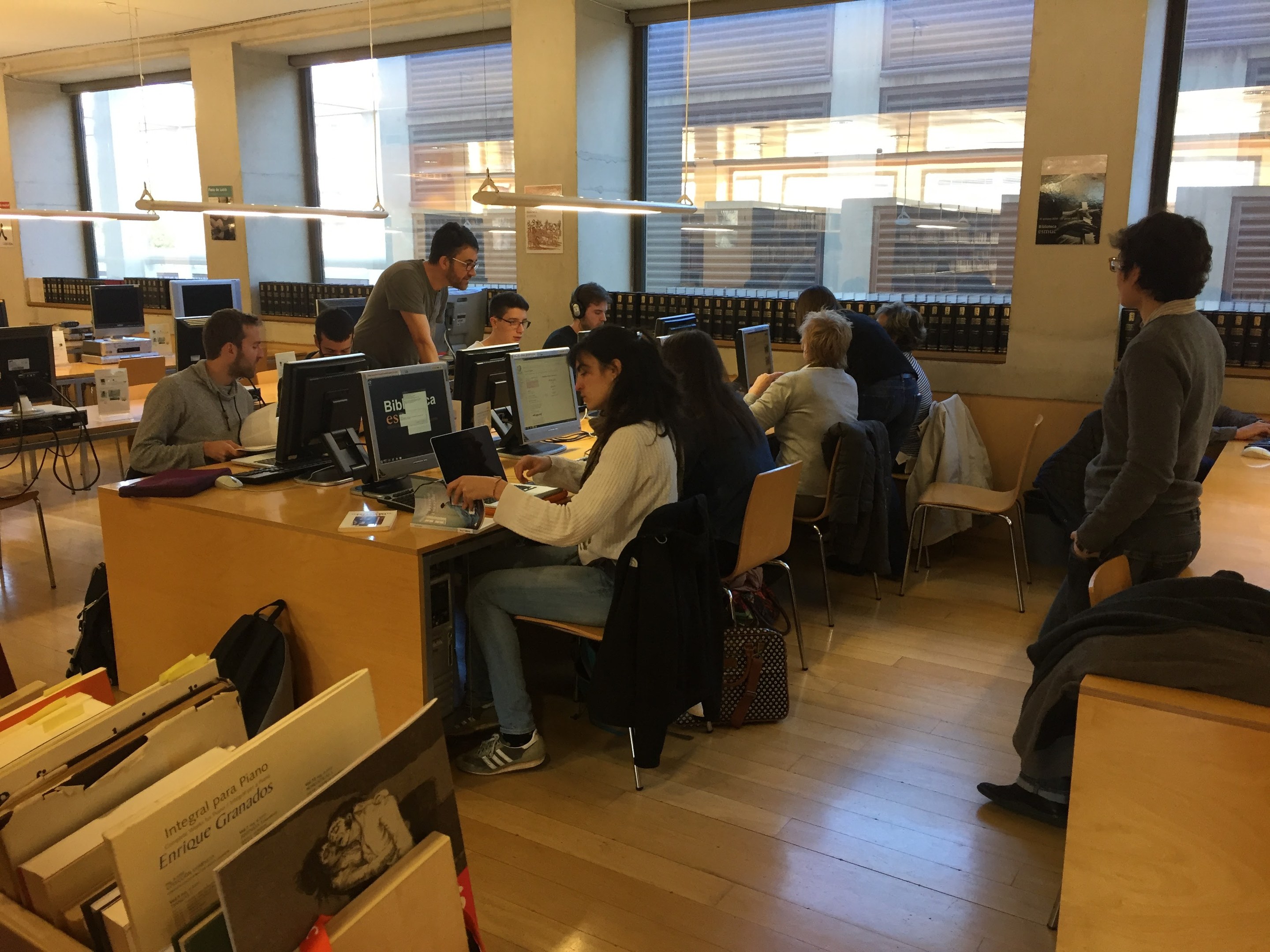 Gruppe von Menschen, die an Computern an Tischen in einer Bibliothek mit Bücherregalen, Monitoren, Tastaturen, Mäusen, Taschen auf dem Boden, Deckenlampen und Glasfenstern im Hintergrund arbeiten.