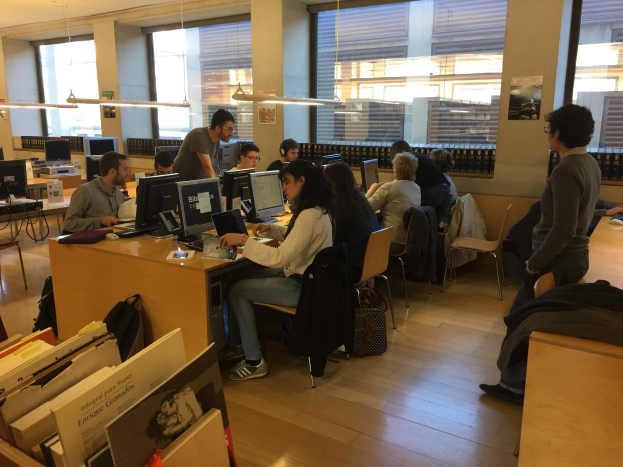 Gruppe von Menschen, die an Computern an Tischen in einer Bibliothek mit Bücherregalen, Monitoren, Tastaturen, Mäusen, Taschen auf dem Boden, Deckenlampen und Glasfenstern im Hintergrund arbeiten.