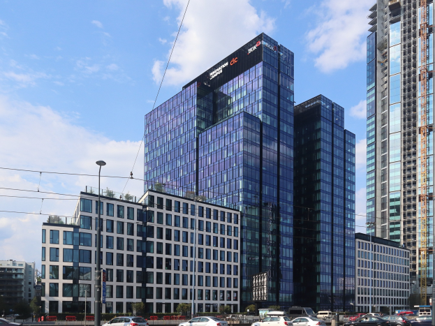 Großes modernes Gebäude mit zahlreichen Fenstern, identifiziert als die Deutsche Bank Hauptstad in Frankfurt, Deutschland, umgeben von städtischer Infrastruktur einschließlich Straßenlaternen, Fahrzeugen, Bäumen und einem bewölkten Himmel.