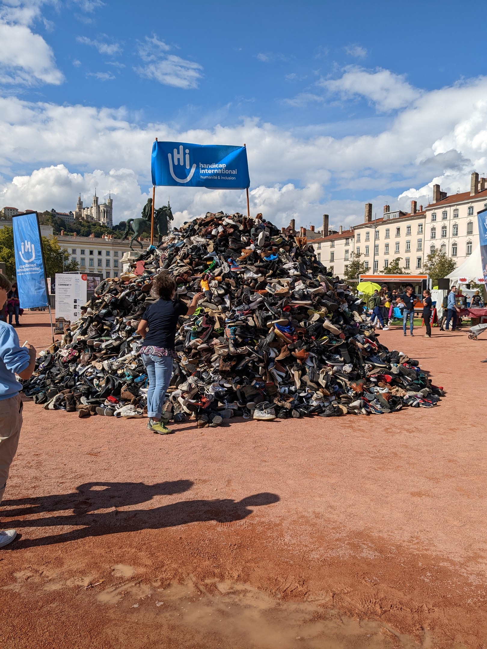 Eine Gruppe von Menschen versammelte sich um einen Haufen Schuhe auf einem Feld mit Gebäuden, Bäumen und einem bewölkten Himmel im Hintergrund bei einem internationalen Schuh-Recycling-Event.