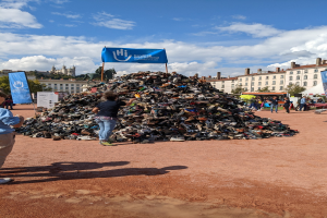 Eine Gruppe von Menschen versammelte sich um einen Haufen Schuhe auf einem Feld mit Gebäuden, Bäumen und einem bewölkten Himmel im Hintergrund bei einem internationalen Schuh-Recycling-Event.