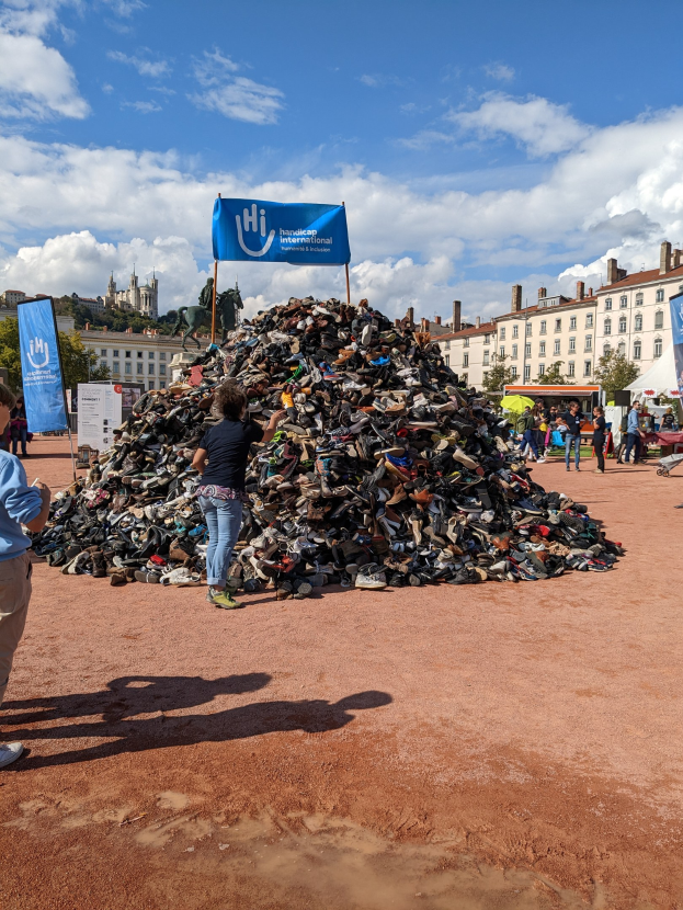 Eine Gruppe von Menschen versammelte sich um einen Haufen Schuhe auf einem Feld mit Gebäuden, Bäumen und einem bewölkten Himmel im Hintergrund bei einem internationalen Schuh-Recycling-Event.