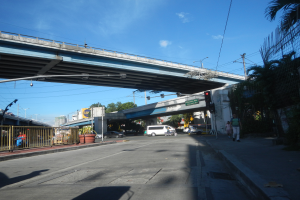 Eine Stadtstraße mit einer Fußgängerbrücke, Fahrzeugen, Fußgängern, Gebäuden und einem bewölkten Himmel im Hintergrund.