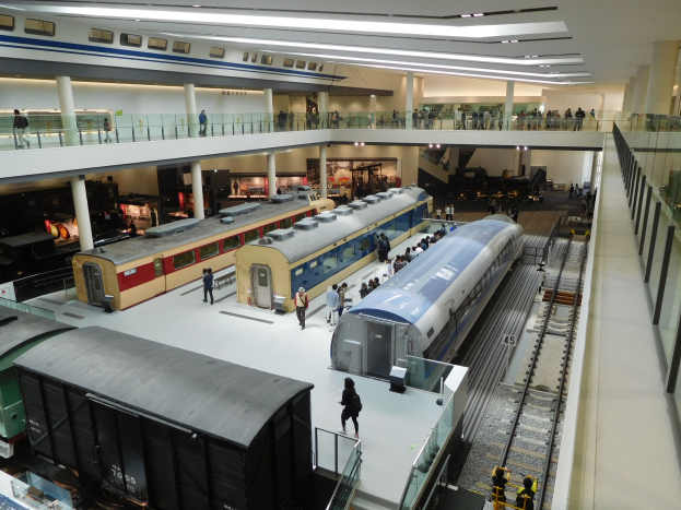 Ein großer Museumsraum mit verschiedenen Zügen in unterschiedlichen Größen und Farben, bei dem Menschen die Ausstellungsstücke beobachten und Deckenlampen den Raum erhellen.