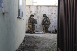 Zwei Soldaten stehen in der Mitte des Bildes, umgeben von Erde, mit einer Wand und einem Fenster auf der linken Seite, einer eisernen Wand auf der rechten Seite und Gebäuden, einem Strommast und einer weiteren Wand im Hintergrund.