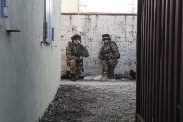 Zwei Soldaten stehen in der Mitte des Bildes, umgeben von Erde, mit einer Wand und einem Fenster auf der linken Seite, einer eisernen Wand auf der rechten Seite und Gebäuden, einem Strommast und einer weiteren Wand im Hintergrund.