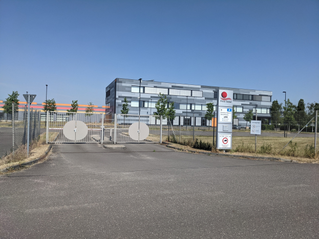 Großes industrielles Gebäude mit der Aufschrift "First Solar" und vielen Fenstern, umgeben von einem Metallzaun, zahlreichen Bäumen, Pflanzen, Gras, Schildern, Straßenlaternen und einem Weg unter einem bewölkten Himmel.