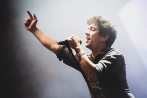 Ein Mann in schwarzem Hemd und Jeans singt leidenschaftlich in ein Mikrofon auf einer hell erleuchteten Bühne, die Augen geschlossen und das Haar stylisch durcheinander, während das Publikum im Hintergrund applaudiert.