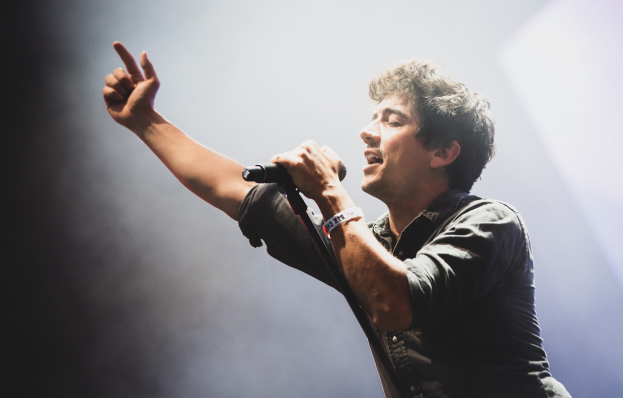 Ein Mann in schwarzem Hemd und Jeans singt leidenschaftlich in ein Mikrofon auf einer hell erleuchteten Bühne, die Augen geschlossen und das Haar stylisch durcheinander, während das Publikum im Hintergrund applaudiert.