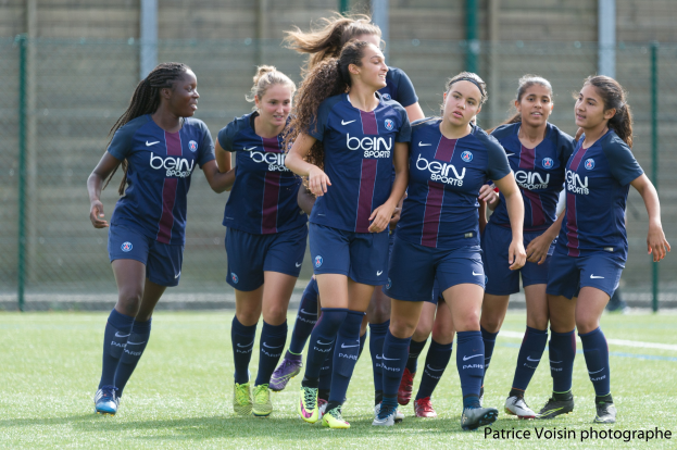Eine Gruppe junger Frauen, die auf einem Rasenfeld Fußball spielen, mit Maschendrahtzaun und einer Wand im Hintergrund, beschriftet mit "Paris Saint-Germain Frauenfußball" in der rechten unteren Ecke.