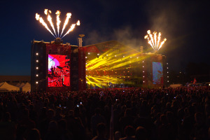 Eine große Menschenmenge auf einem Musikfestival mit einer Bühne, auf der Künstler und helle Lichter zu sehen sind, Zelte links daneben und Feuerwerk am dunklen Himmel.