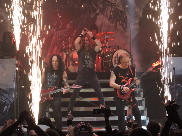 Eine Gruppe von Menschen, die auf einer Bühne bei einem Konzert spielen, wobei einige Gitarren halten und einer ein Mikrofon hält, während ein anderer hinter ihnen Schlagzeug spielt; Lautsprecher sind auf der linken Seite zu sehen, Treppen führen zur Bühne hinauf und eine Menge mit Handys ist unten zu sehen.