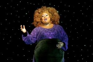 Eine Frau in einem lila Kleid steht auf einer Bühne, beleuchtet von Hintergrundlichtern und scheint mitten in einer Vorstellung zu sein.