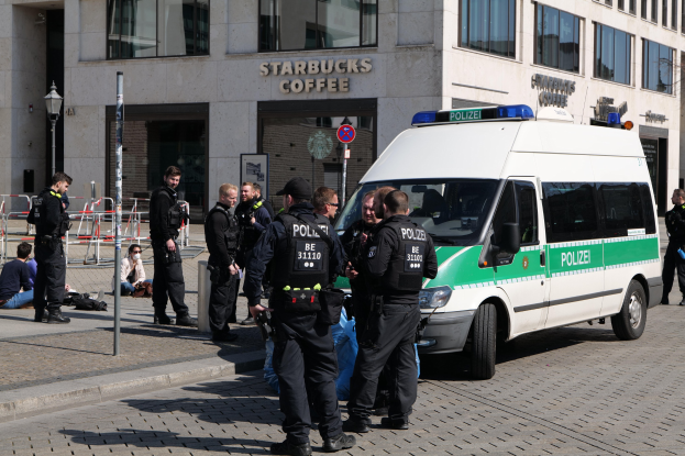 Eine Gruppe von Polizeibeamten steht vor einem Starbucks-Café, rechts davon parkt ein Van und links stehen und sitzen einige Menschen. Im Hintergrund ist ein Gebäude mit Fenstern, ein Schild, ein Laternenmast und ein Zaun zu sehen.