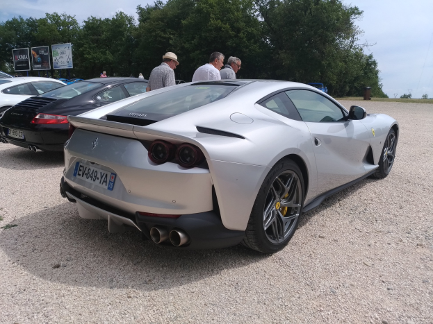 Roter Ferrari 812 Superfast auf dem Goodwood Festival of Speed umgeben von Menschen mit Kappen, Bäumen, Texttafeln und einem bewölkten Himmel im Hintergrund.