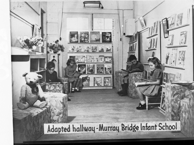 Gruppe von Kindern auf Stühlen in einem bibliotheksähnlichen Raum mit Bücherregalen, ein Teddybär links, eine Blumenvase auf dem Tisch, gerahmte Gegenstände an der Wand und Text unten "Angepasster Flur Murray Bridge Kindergarten"
