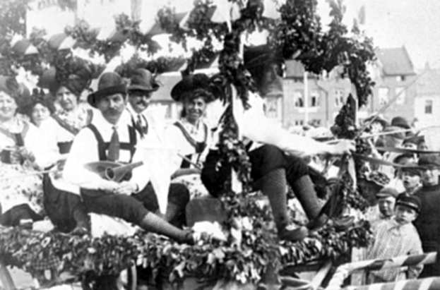 Eine Gruppe von Menschen in traditioneller bayrischer Tracht fährt auf einemblattgeschmückten Festwagen in einem Umzug mit, einige spielen Musikinstrumente, mit Gebäuden und einem klaren Himmel im Hintergrund, dargestellt in Schwarz-Weiß.