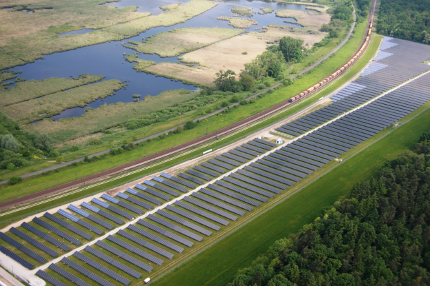 Luftaufnahme eines Solarparks mit Panelen, umgeben von Bäumen, Gras, Pflanzen und Wasser, mit einem Zug auf einer nahen Bahnstrecke.