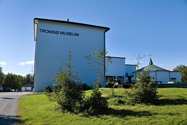Das Tromsø-Museum in Norwegen, mit einem weißeem Geböude mit grünem Rasen, Böumen und parkenden Fahrzeugen unter einem teilweise bewölktem Himmel.