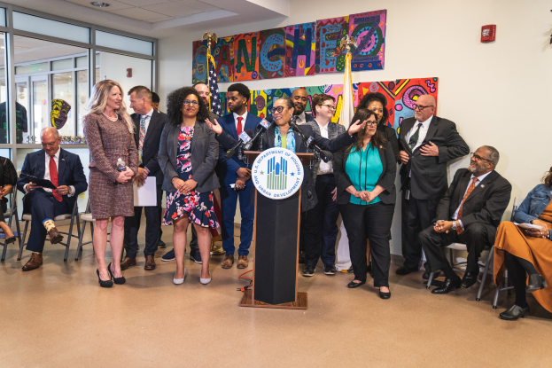 Eine Gruppe von Menschen steht vor einem Podium mit einem Mikrofon und einer Tafel mit der Aufschrift "Sancho-Stadtrat tritt dem Stadtrat bei", während andere im Hintergrund sitzen und stehen, Flaggen, eine Tafel, ein Feuerlöscher und Deckenleuchten an der Rückwand.