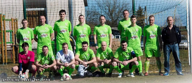 Eine Gruppe von Männern in grünen T-Shirts steht auf einem Fußballfeld, einige sind in der Hocke, mit einem Ball und einem Torpfosten im Hintergrund; der Hintergrund enthält Bäume, Gebäude und Fahrzeuge.