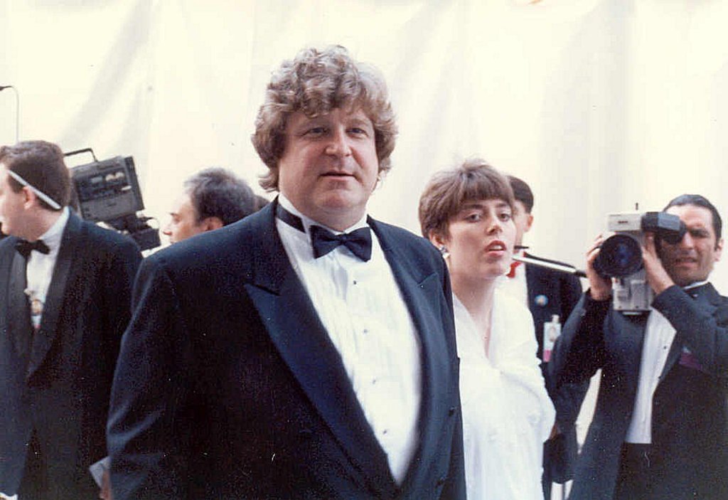 Mehrere Personen stehen, eine Person rechts hält eine Kamera und filmt, links eine Kamera und ein Kabel sowie ein großer weißer Vorhang im Hintergrund.