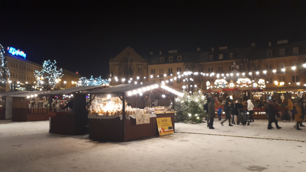 Ein belebter Weihnachtsmarkt an einem verschneiten Abend mit Menschen, Buden mit Lichtern, Pflanzen, Bäumen, Gebäuden und Schildern unter einem bewölkten Himmel.