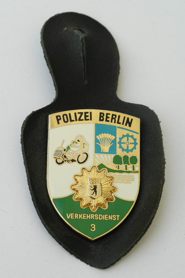 Ein schildförmiges Polizeibadge mit "Polizei Berlin" in schwarzer fetter Schrift auf einem blauen Hintergrund mit einem weißen Stern und Rahmen, das auf einer Oberfläche ruht.