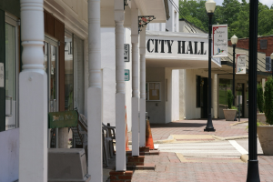 Stadtverwaltung mit umgebender Infrastruktur, einschließlich Straßenlaternen, Schildern, Fahrzeugen, Verkehrskegeln, Stühlen, Zimmerpflanzen, Bäumen und einem bewölkten Himmel.