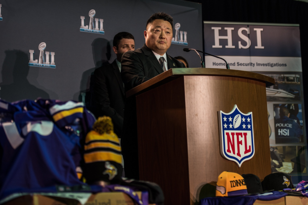 Ein Mann in einem schwarzen Blazer, einem wei├čem Hemd und einer schwarzen Krawatte spricht an einem Podium mit Mikrofon und Logo vor einer Menge. Blaue und gelbe Merchandising-Artikel und ein "Super Bowl LII"-Banner sind im Hintergrund zu sehen.