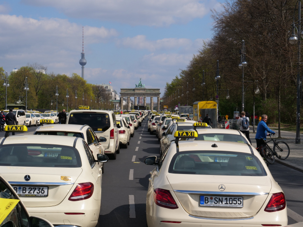 Eine lange Reihe von Taxis steht an einer belebten Straße in Berlin, Deutschland, mit Fahrradfahrern und Förderern auf dem Gehweg, flankiert von Laternen und Bäumen, und Gebäuden, einem Bogen und einem Turm im Hintergrund bei bewölktem Himmel.