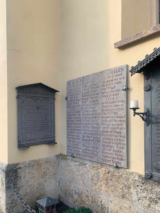 Ein Gebäude mit einer Holocaust-Gedenktafel, einem Fenster, einer Tür, einer äußeren Beleuchtung, einer Kette und umgebenden Pflanzen.