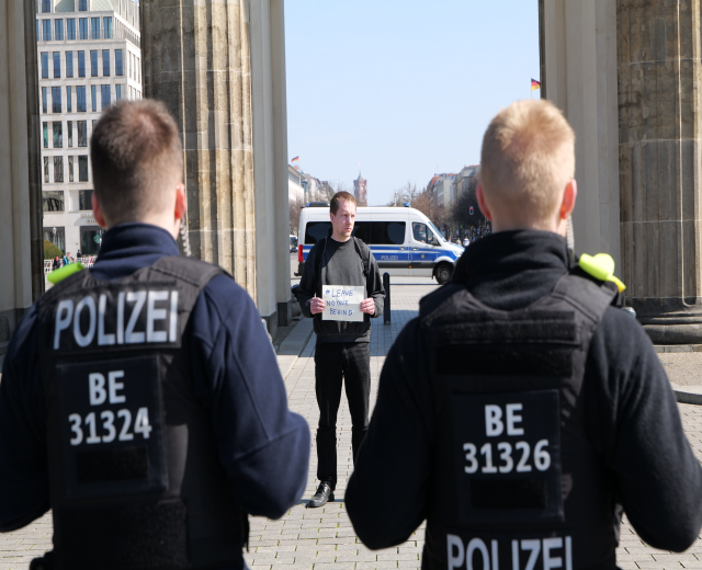 Drei Polizeibeamte stehen vor einem Gebäude, wobei ein Mann in der Mitte ein Papier hält, flankiert von Säulen und vor dem Hintergrund von Fahrzeugen, Menschen, Bäumen, Gebäuden und Himmel.
