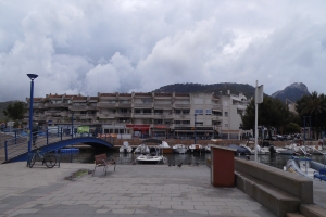 Ein Hafen mit Booten im Wasser, eine Brücke mit Geländern, Bänken, Laternenpfählen, Bäumen, Gebäuden mit Fenstern, Hügeln im Hintergrund und einem Himmel mit Wolken.