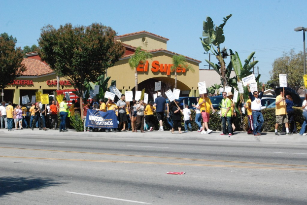 Gruppe von Menschen mit Schildern, die gehen, mit kleinen Büschen, einer Fahne, einem Gebäude mit Namensschildern und Autos im Hintergrund.
