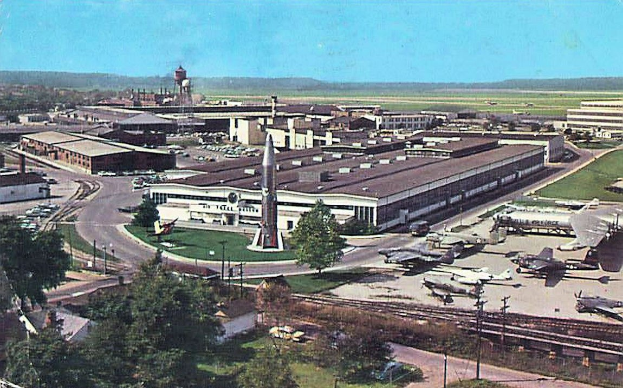 Altes Schwarz-Weiß-Foto eines Flughafens mit geparkten Flugzeugen, Gebäuden, Bäumen, Straßen, Fahrzeugen, Strommasten, Gras, Bahnschienen, Türmen, Hügeln und einem klaren Himmel mit Text am unteren Rand.