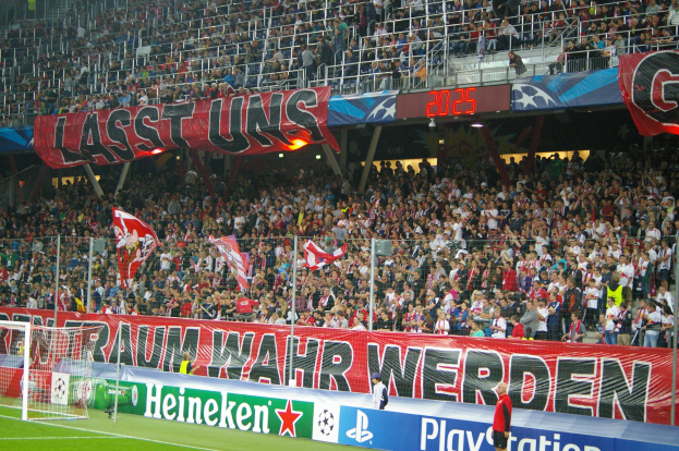Fußballspiel in einem Stadion mit großer Zuschauermenge, saftig grünem Rasen, einem Tor auf der linken Seite, Bodenbanner, Menschen mit Fahnen und Hintergrundgeländer, Tafeln, Lampen und andere Gegenstände.