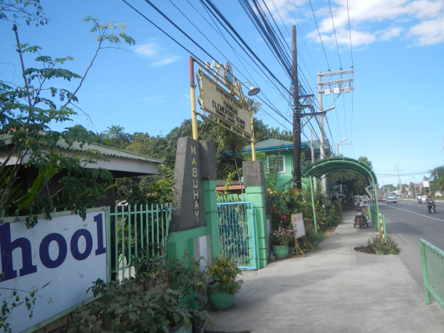 Einfahrt zu einer Schule mit einem "Schule"-Schild, Fahrzeugen auf der Straße, einem Bogen, einem Tor, einer Namensplakette, Pflanzen, Bäumen, Strommasten mit Drähten und einem bewölkten Himmel im Hintergrund.