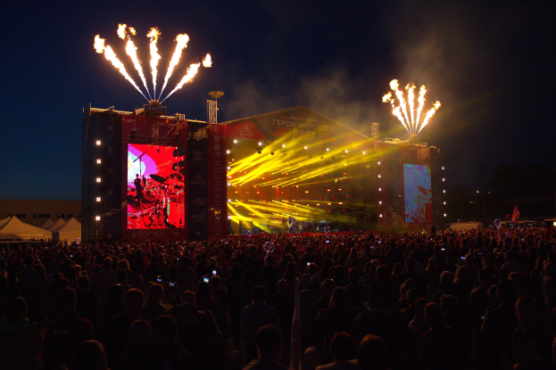 Eine große Menschenmenge auf einem Musikfestival mit einer Bühne, auf der Künstler unter hellen Scheinwerfern spielen, umgeben von Bannern, Zelten links daneben und einem Feuerwerk, das den dunklen Himmel erhellt.