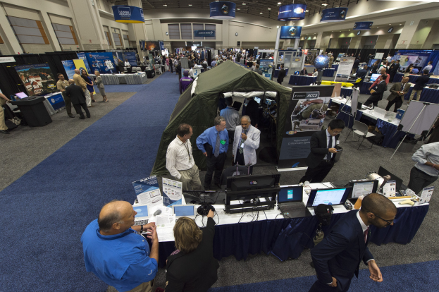 Eine große Gruppe von Menschen auf einer Karriere-Messe um einen Tisch mit Laptops, Monitoren und Papieren versammelt, mit einem Zelt, Informationsplakaten, Deckenlampen und Fenstern im Hintergrund.