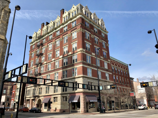 Historisches Backsteingebäude in Apartments umgewandelt an einer Straßenecke, mit Fahrzeugen, Passanten, Straßenmöblierung und Bäumen im Hintergrund.