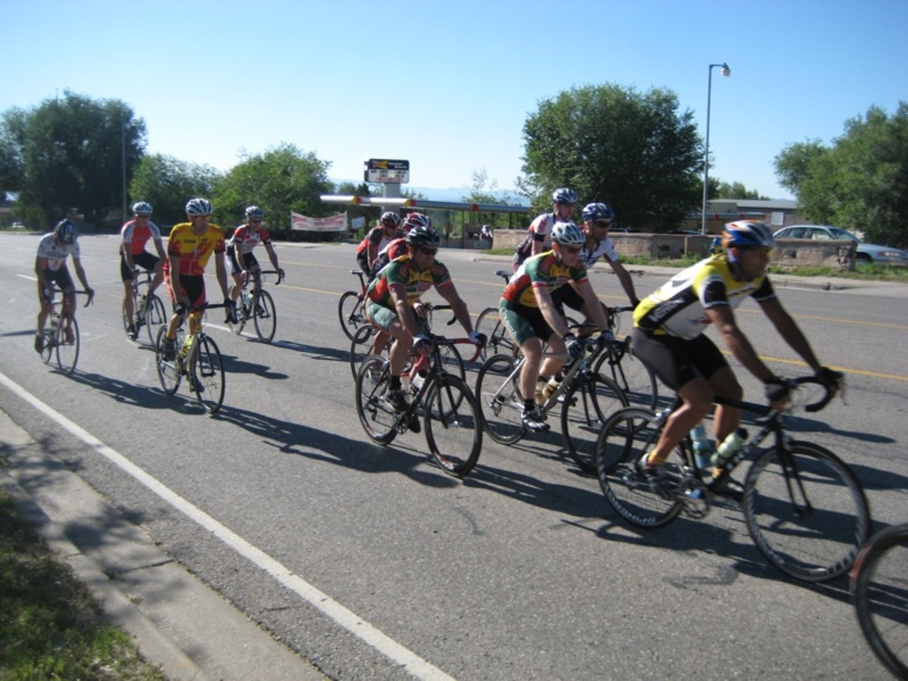 Eine Gruppe von Menschen mit Helmen und Schutzbrillen auf Fahrr√§dern auf einer Stra√Ÿe, mit zwei Pfosten, einem mit einer Lampe, einem Fahrzeug, einem Geb√§ude, einem Schuppen mit einer Tafel √ľber ihm, B√§umen, Gras und dem Himmel im Hintergrund.