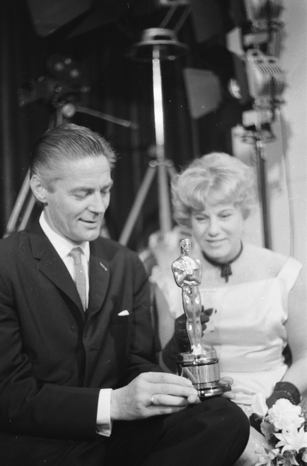 Ein Schwarz-Wei├č-Bild eines formell gekleideten Mannes mit einer Oscar-Statue und einer Frau mit einem Blumenstrau├čus, die nebeneinander sitzen und im Hintergrund beleuchtet sind.