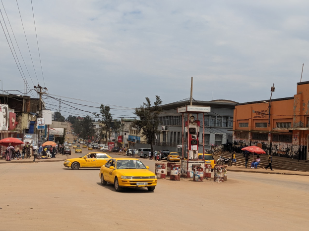 Eine belebte Stadtstraße mit einem gelben Taxi, das fährt, Passanten mit Schirmen auf den Gehwegen, Gebäude mit Fenstern und Strommasten mit Drähten unter einem bewölkten Himmel.