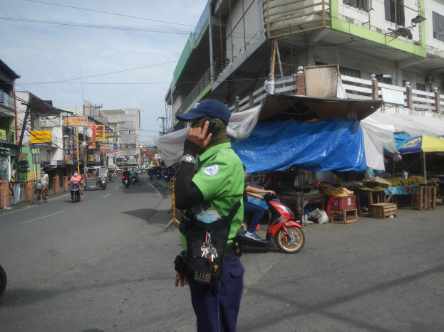 Ein Polizeibeamter in einer grünen und blauen Uniform und einer blauen Mütze steht auf der Seite einer Straße und spricht auf einem Handy, mit Fahrzeugen, die hinter ihm fahren, Ständen mit verschiedenen Gegenständen zu seiner Rechten und einem bewölkten Himmel mit einem Gebäude mit Fenstern im Hintergrund.