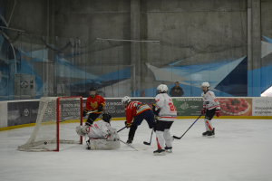 Gruppe von Menschen beim Eishockey auf einer Indoor-Eisfläche, die Helme tragen und Eishockeyschläger halten, mit einem Torpfosten links und Bannern im Hintergrund.