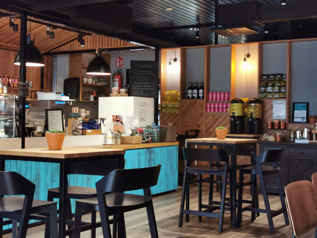 Innenraum eines Restaurants mit Holztischen und -stühlen, einem Blumentopf auf einem Tisch, einer Schiefertafel, einem Kühlschrank, einem Feuerlöscher, Regalen mit Flaschen, Schränken mit Gegenständen, einem gerahmten Foto an der Wand, Deckenleuchten und Lampen.