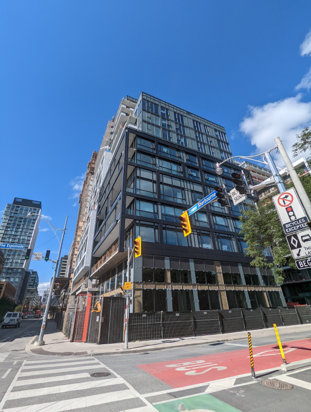 Eine belebte Stadtstraße mit Fahrzeugen, Ampeln, Schildern, Bäumen und einem Zaun, mit einem hohen Apartment/Wohnhausgebäude, das als Verkaufsobjekt an 541 East 57th Street, Unit 541, gekennzeichnet ist.