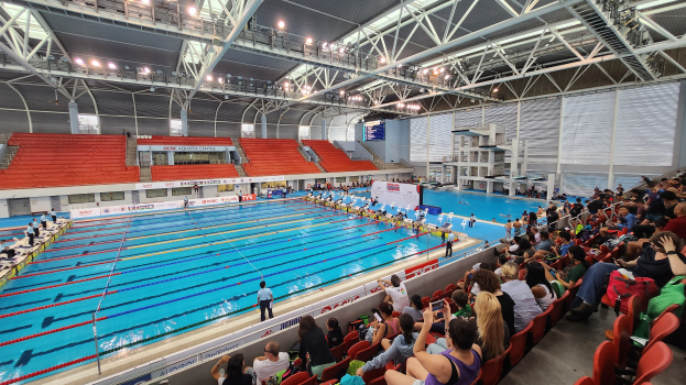 Großer Hallenbad mit vielen Menschen auf Stühlen drumherum und einigen im Wasser Stehenden, mit hoher Decke mit Lampen und Eisenstangen und Werbetafeln im Hintergrund.