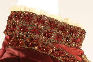 Nahaufnahme eines roten und goldenen Kleides mit detaillierter Spitzenverzierung auf einem Schaufensterpuppen gegen einen weißen Hintergrund.